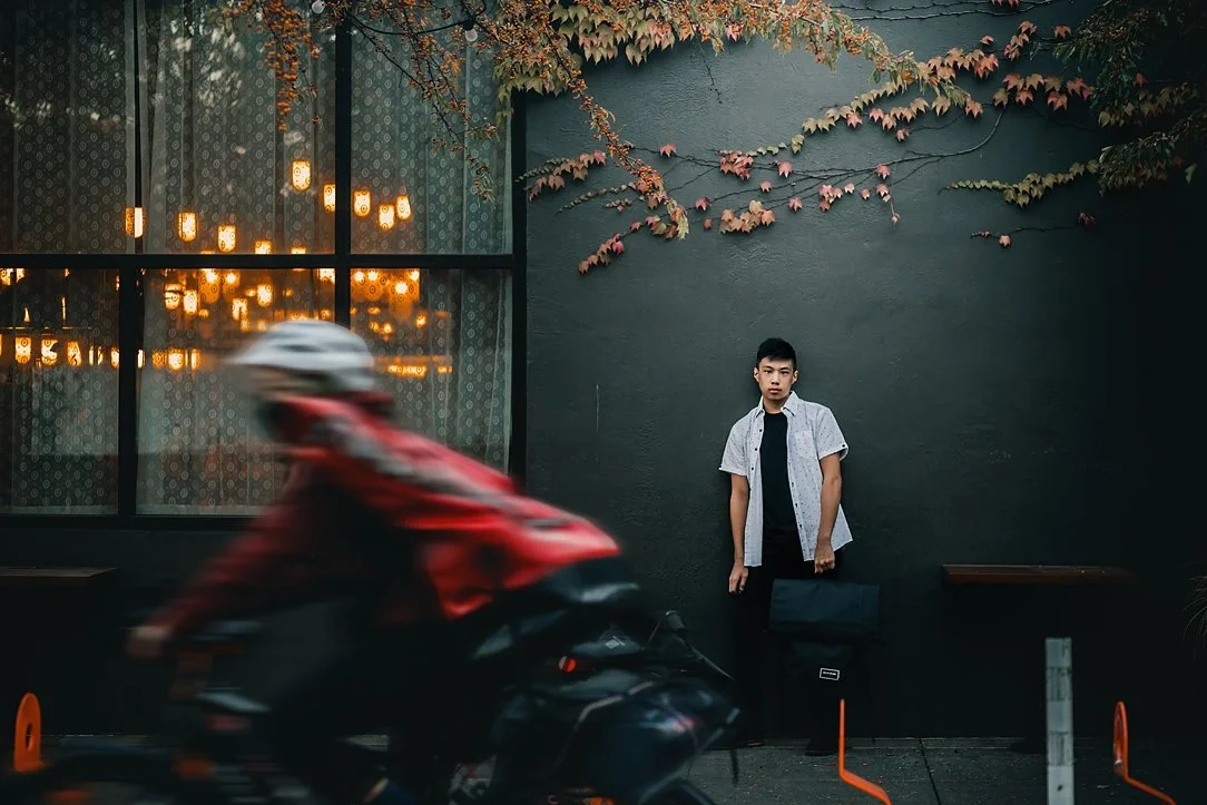 A high school senior in a white button-down shirt holding a backpack stands against a dark grey wall with ivy in Portland, with a blurred bicyclist in motion in the foreground.