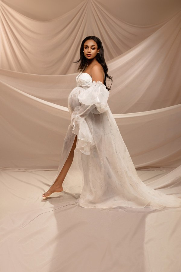 A pregnant woman with long dark hair walking in a studio while wearing a flowing white off-the-shoulder maternity gown with floral details, set against draped cream fabric backdrops.