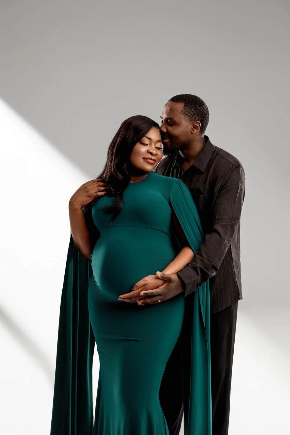 Fine-art studio maternity portrait of an expectant couple posing against a light grey backdrop in West Linn, Oregon, with the mother in an emerald green gown cradling her baby bump.