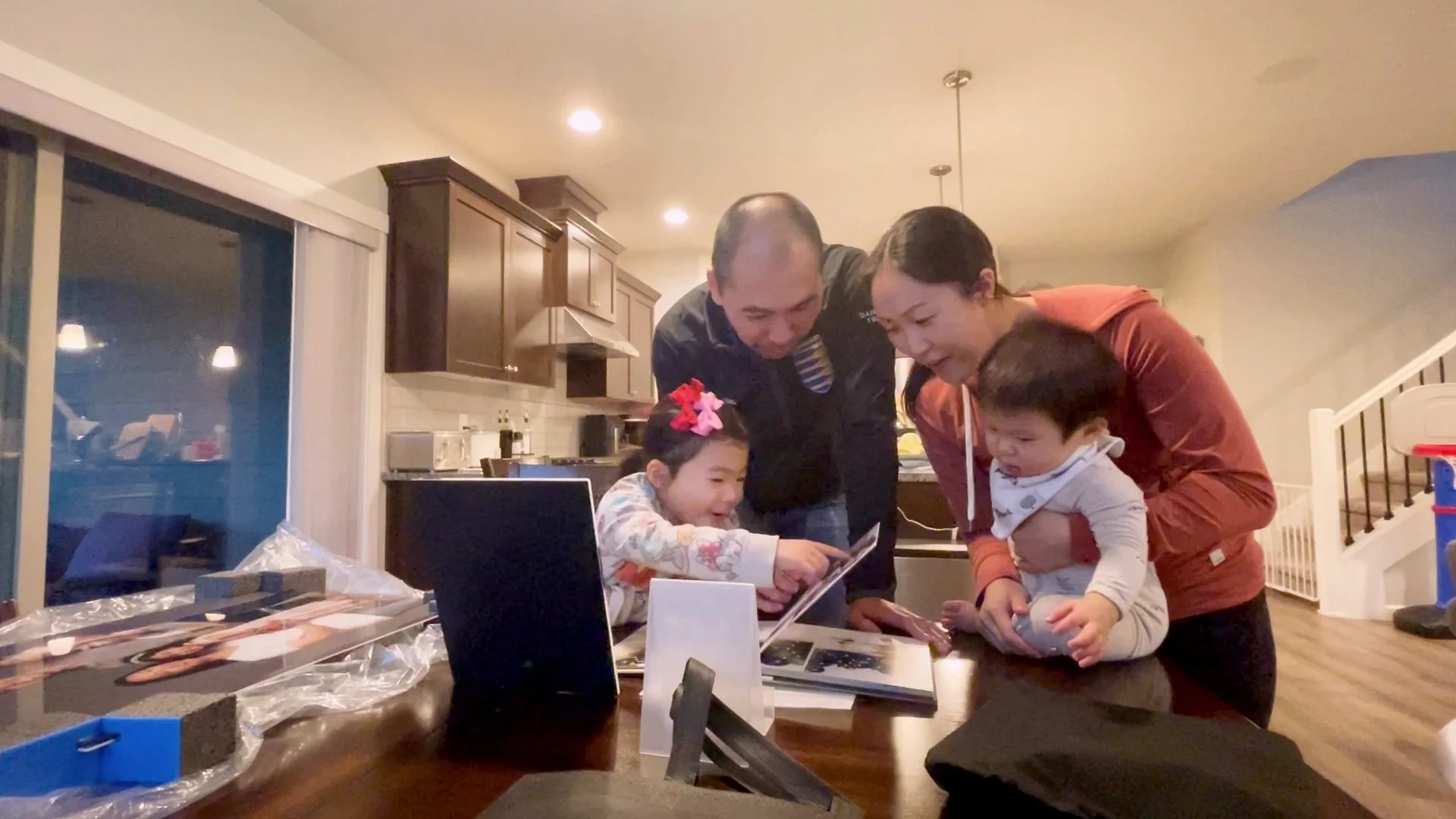 A heartwarming candid moment of a family of four—mother, father, and two young children—gathered around a table to view their new professional photo album. A young girl points excitedly at the pages while the parents smile, showcasing the gallery rev