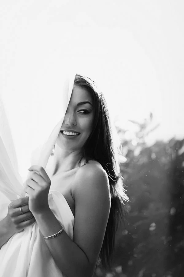 A black and white portrait of a smiling woman partially veiled by a white fabric sheet, standing outdoors with soft, natural backlighting and a blurred foliage background.