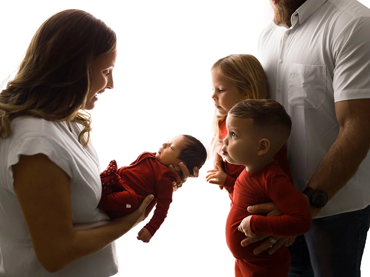 A professional studio photograph of a family of five. The mother is on the left, smiling at her sleeping newborn who is dressed in a deep red ribbed outfit. Two older children—a girl and a boy—stand in the center looking at the baby, also wearing mat