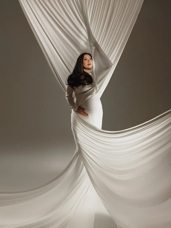 A pregnant woman with long dark hair wearing a fitted white maternity gown, standing as long lengths of white silk fabric are tossed around her in a studio.