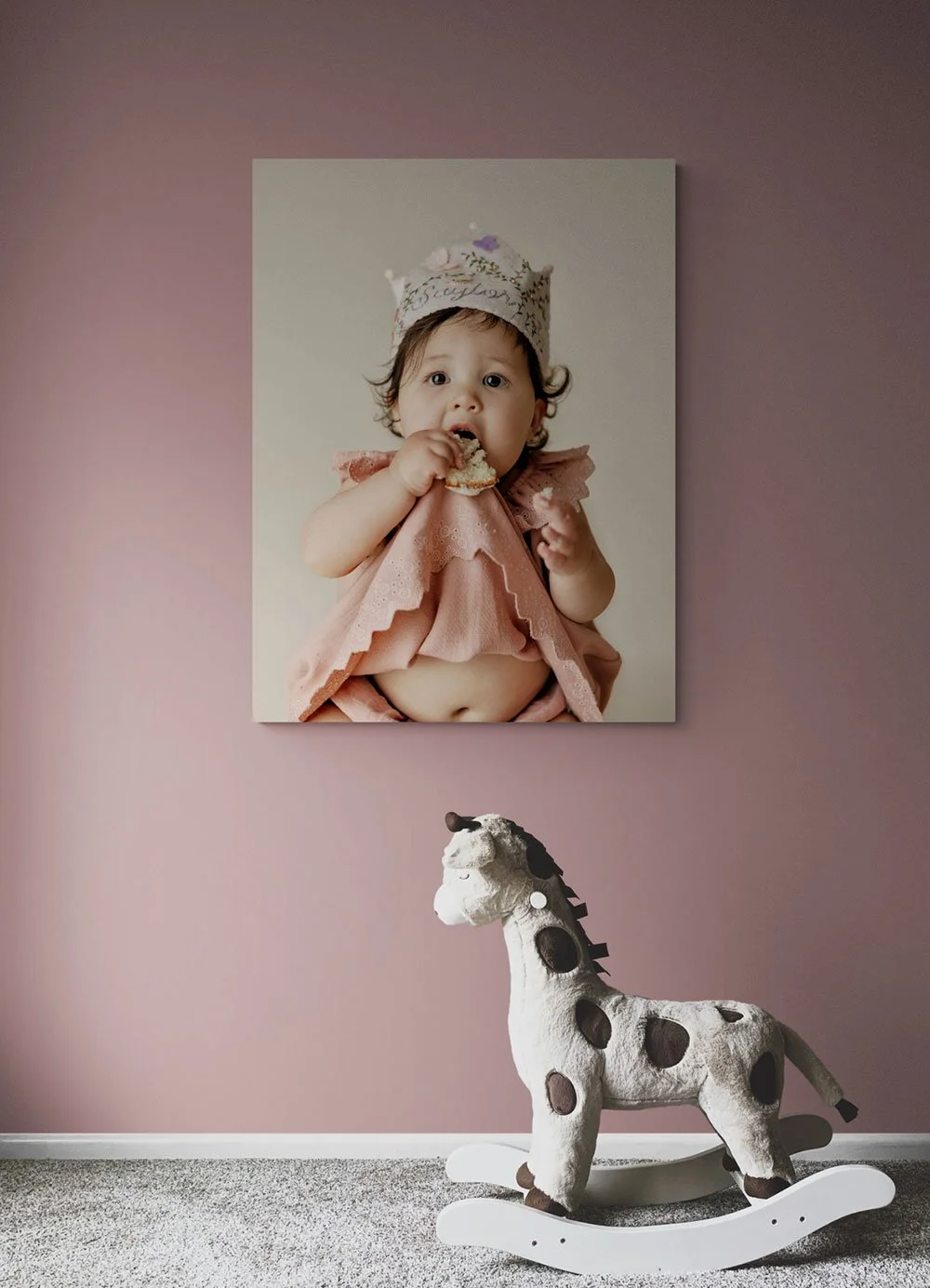 Vertical first birthday milestone canvas portrait mockup hanging on a dusty rose wall above a plush giraffe rocking horse.