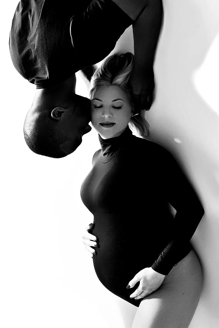 Sunnymelon Photography black and white studio portrait of a maternity couple; the woman is wearing a black turtleneck bodysuit and embracing her pregnant belly while the man kisses her cheek from above.