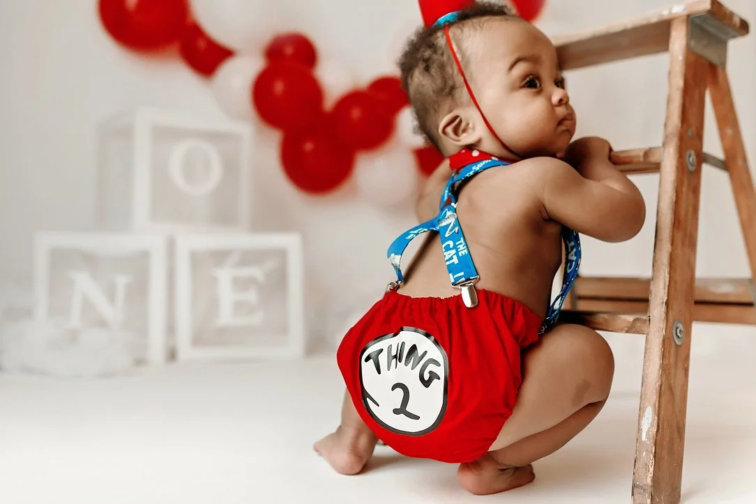 This adorable milestone portrait captures a candid moment from a "Thing 2" themed first birthday session. The baby is dressed in custom Dr. Seuss-inspired attire, featuring a red diaper cover with the "Thing 2" emblem and blue graphic suspenders. Pos