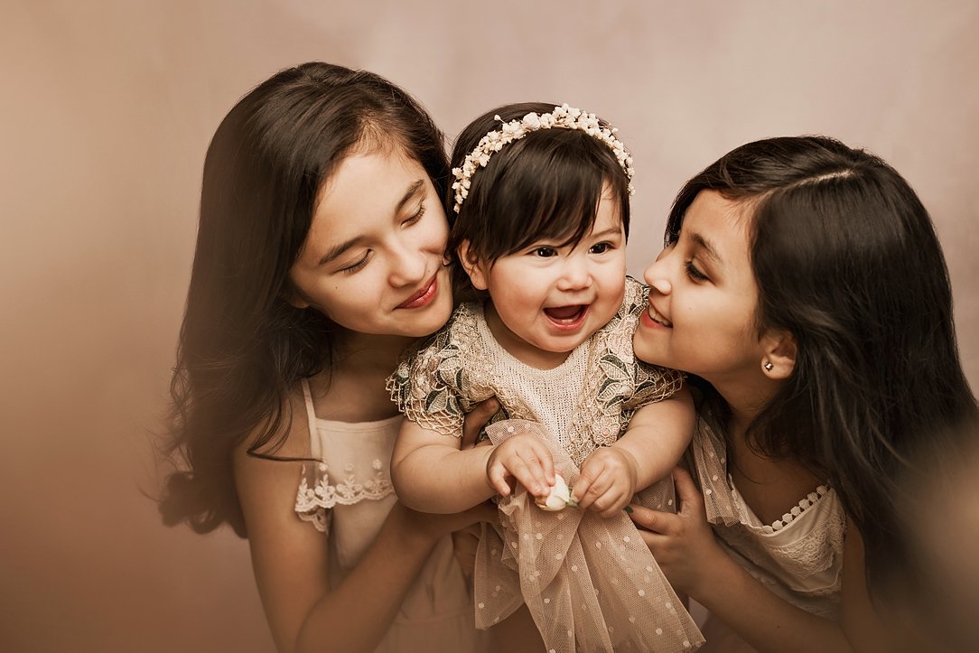 A studio portrait of three smiling young girls; two older sisters wearing matching cream lace dresses lean in to cuddle a laughing baby sister who has a flower crown and a dotted tulle outfit, against a warm, neutral background.