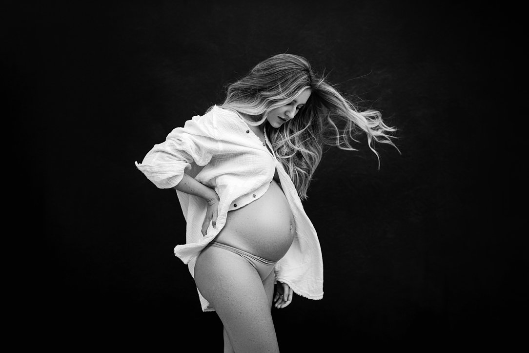 Black and white Sunnymelon maternity portrait of a pregnant woman with windblown hair, wearing an unbuttoned white shirt against a dark studio background.