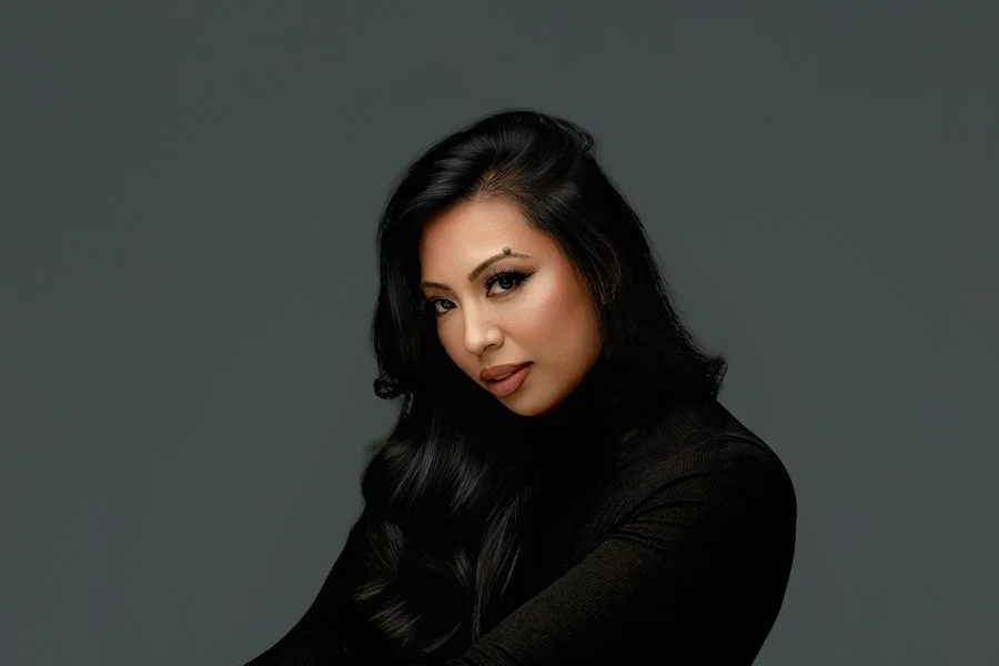 A head-and-shoulders fine art portrait of a woman with long wavy black hair, wearing a black turtleneck and posing against a minimalist grey studio background.