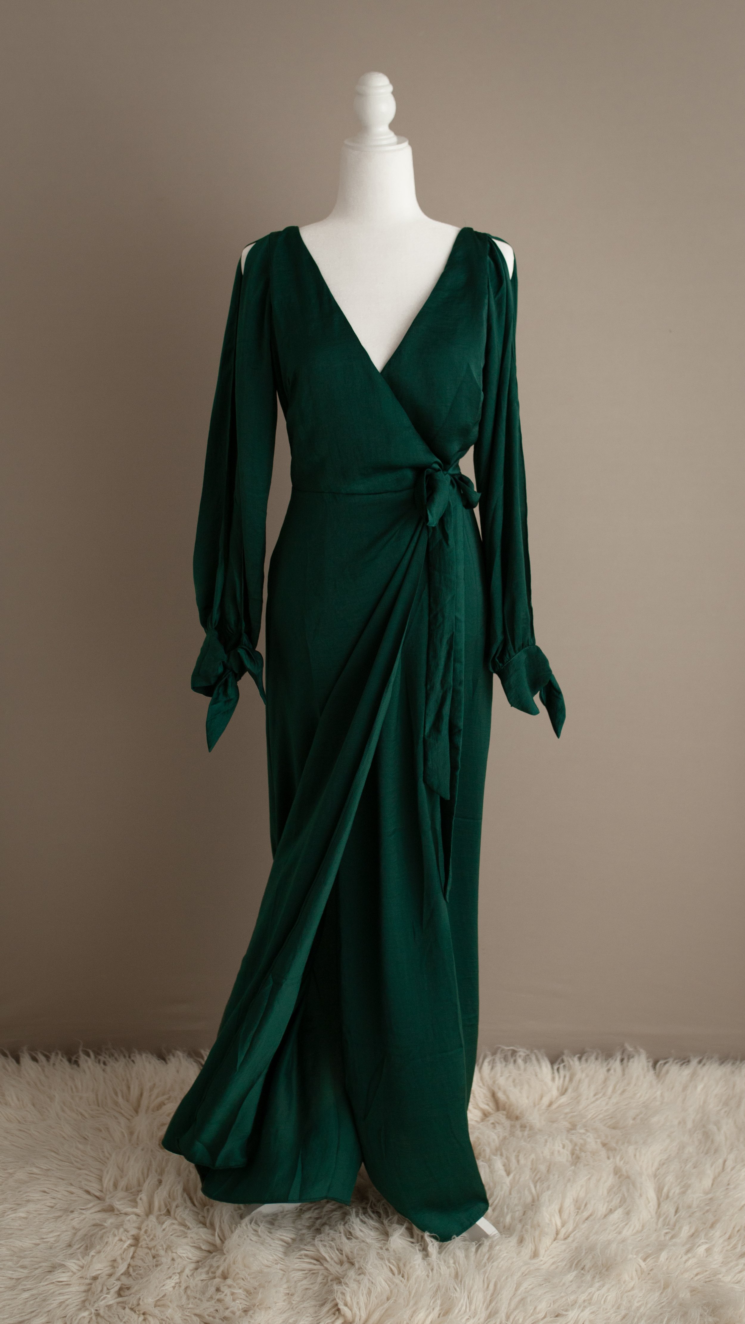 Emerald green satin long-sleeve wrap dress with a V-neck and shoulder cutouts, tied at the waist, displayed on a white mannequin on a fluffy rug.