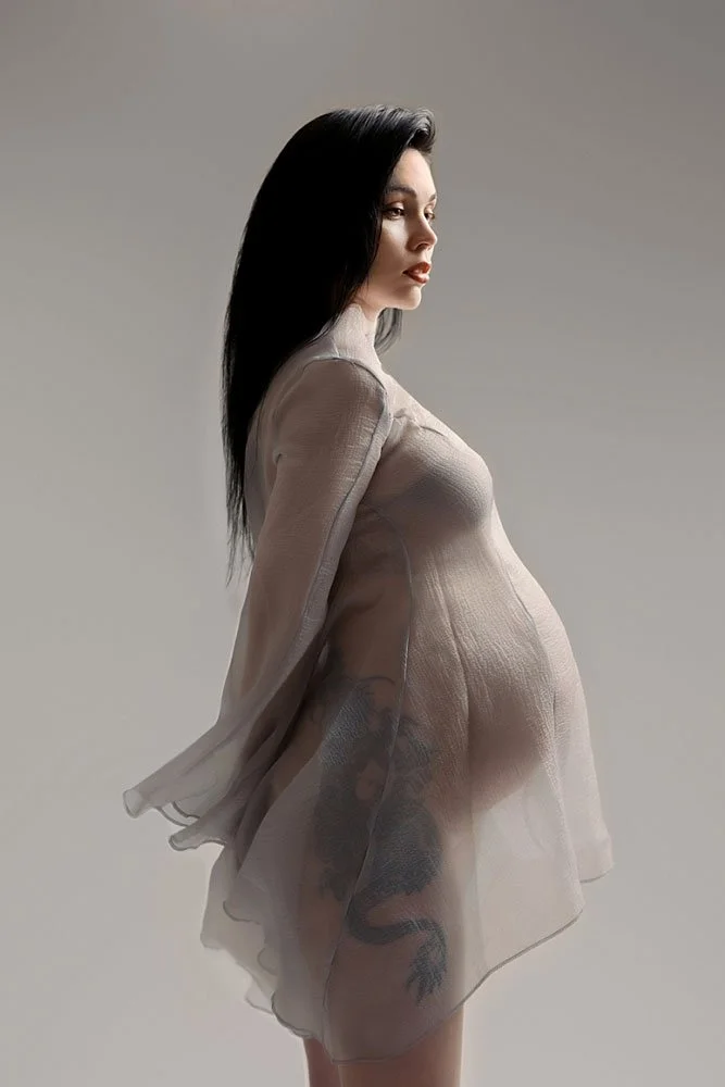 Sunnymelon Photography fine art maternity portrait of an expectant mother in a flowy, sheer grey gown showcasing her baby bump and large leg tattoo in profile against a light grey background.