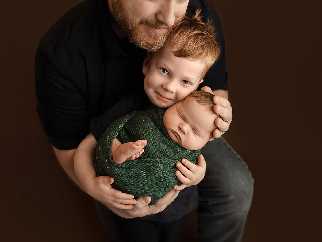 A soulful studio newborn portrait featuring a father holding his young son, who is cuddling a sleeping newborn baby. The newborn is snugly wrapped in a textured forest green swaddle. The trio is posed against a solid dark brown background, creating a