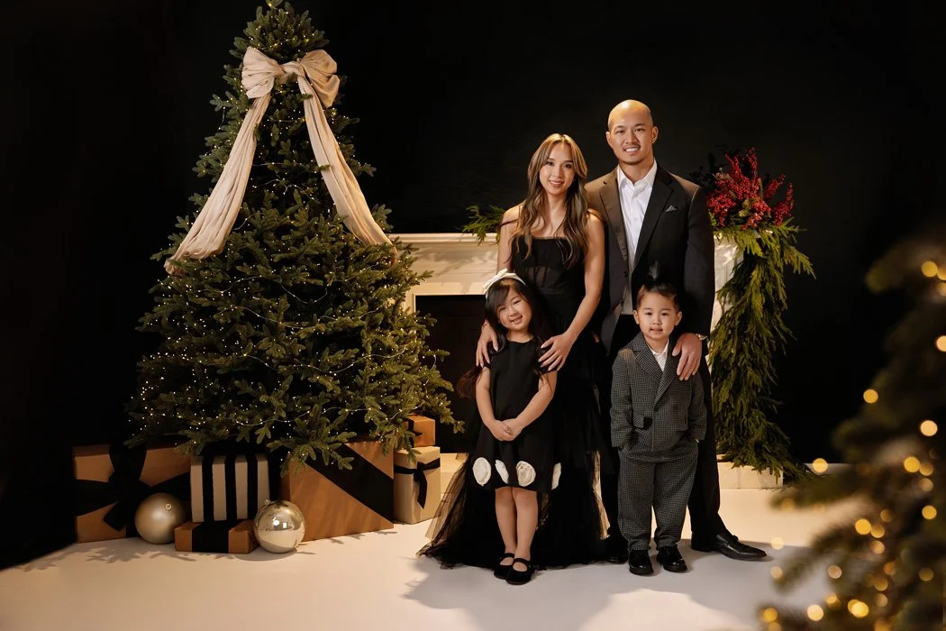 A formal studio portrait of a family of four posing next to a decorated Christmas tree in Portland. The parents and children are dressed in elegant black and grey attire against a classic holiday backdrop.