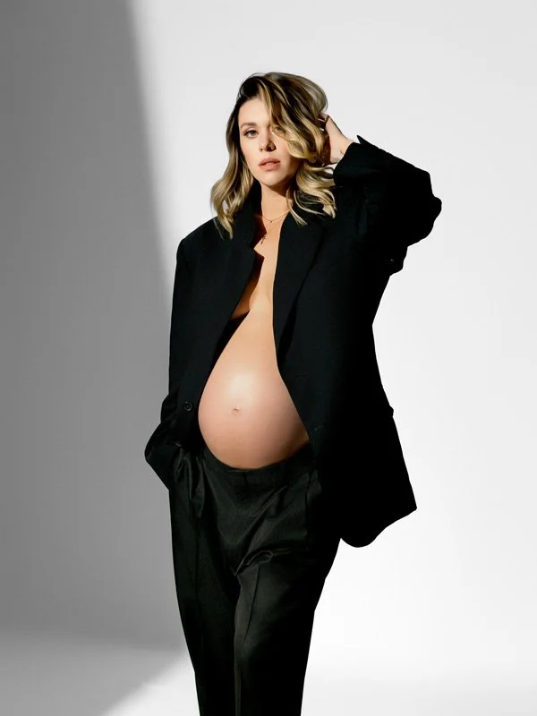 A pregnant woman with blonde wavy hair posing in a studio wearing an open oversized black blazer and matching trousers, highlighting her bare baby bump against a bright white background.