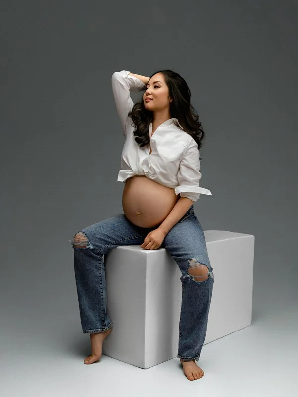 A pregnant woman with long dark hair sitting on a white studio block wearing a tied white button-down shirt and distressed blue jeans against a gray backdrop.