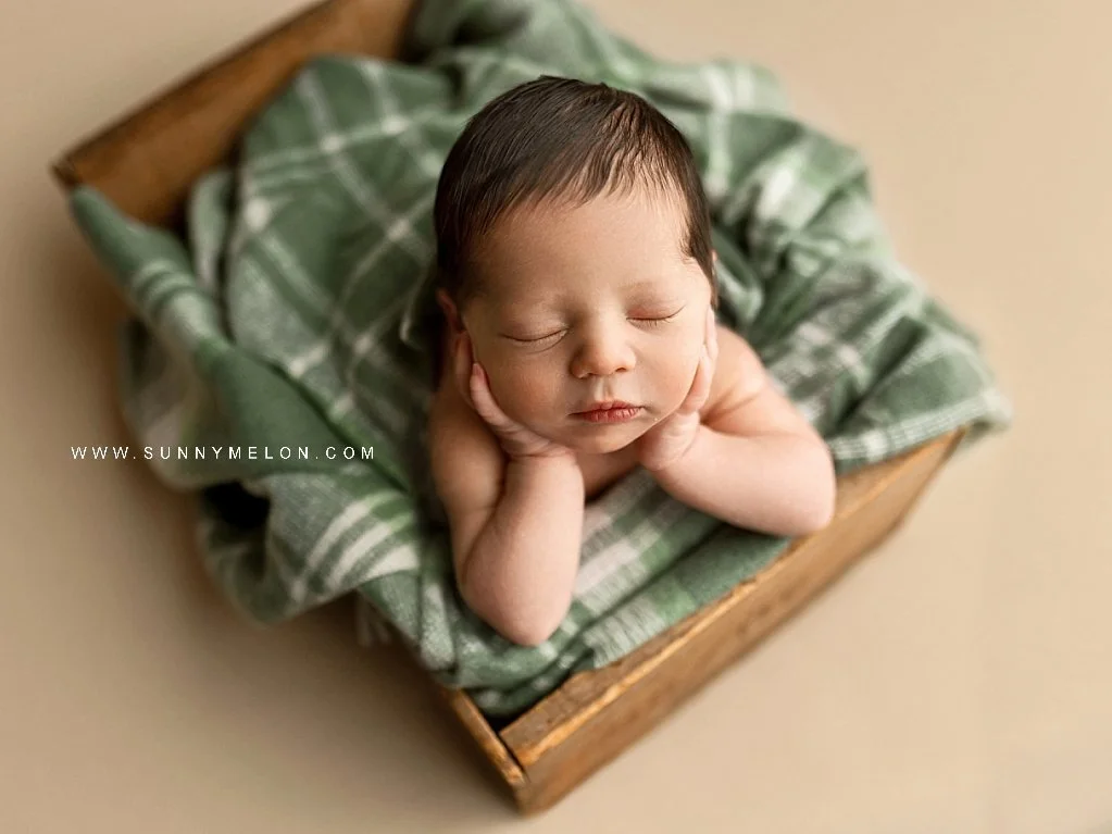 Sleeping newborn baby in a "froggie pose" resting in a rustic wooden crate with a green plaid blanket.