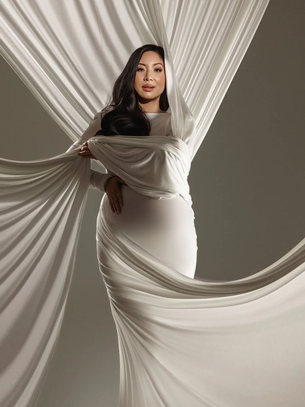 Fine-art studio maternity portrait of a pregnant woman in a white gown artistically draped in flowing white fabric against a grey backdrop in West Linn, Oregon.