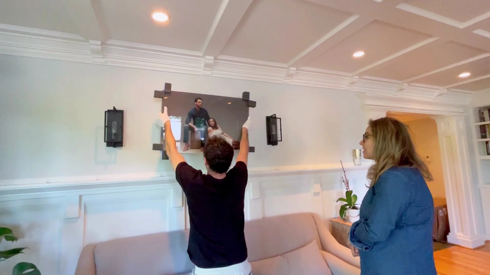 A man wearing white handling gloves carefully installs a large, custom-framed family portrait on a client's living room wall. A woman watches as her new heirloom artwork is perfectly placed above the mantle, showcasing the premium end-to-end service 