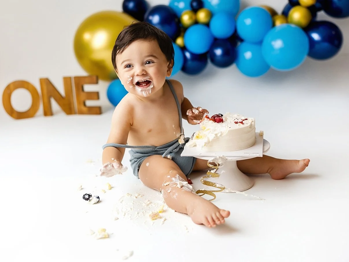 This joyful milestone portrait features a baby boy celebrating his first birthday with a classic cake smash session. The infant is captured mid-laugh, covered in white frosting while sitting next to a partially eaten white cake. The set is styled wit