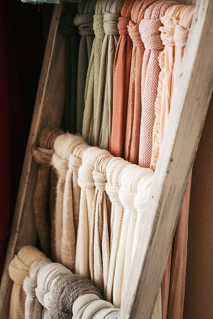This detail shot highlights a beautifully organized collection of high-quality newborn swaddle wraps displayed on a rustic wooden ladder. The wraps feature a variety of textures, including knits and soft jerseys, in a cohesive earth-toned color palet