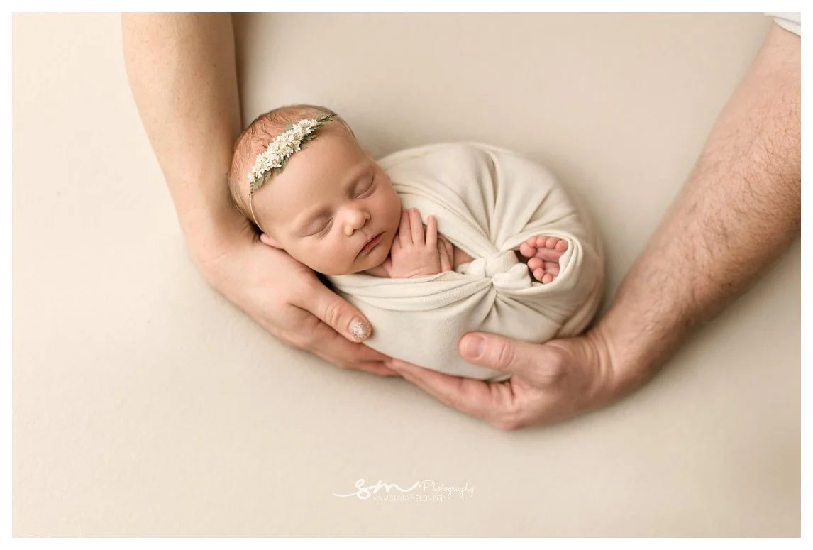 An overhead studio newborn portrait against a cream background showing a swaddled sleeping baby girl with a floral headband being cradled gently in her parents' hands.