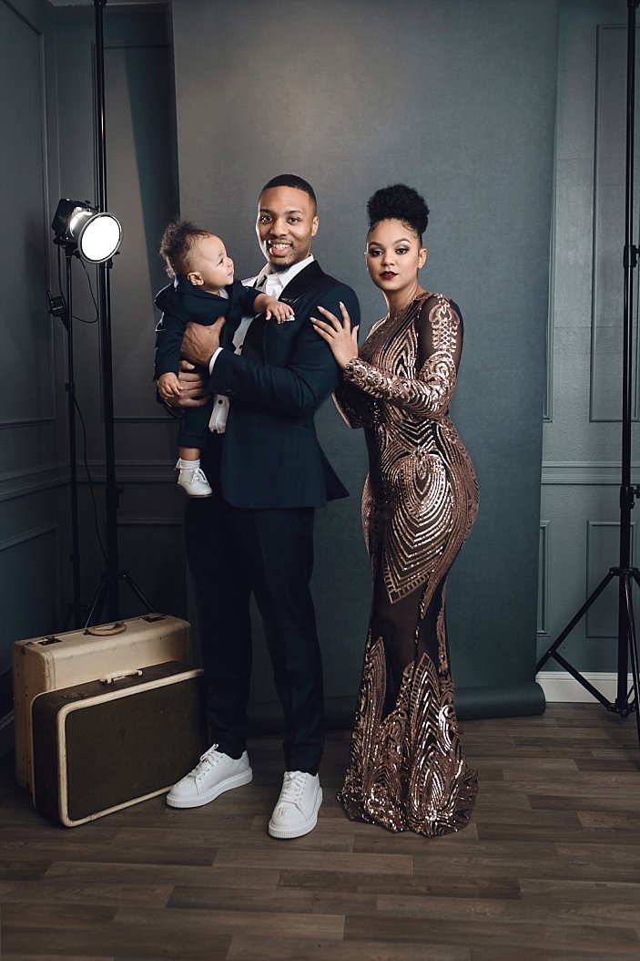 This sophisticated image captures a formal family portrait session in a professional studio setting. The subjects—Damian Lillard, Kayla in an elegant evening gown, and their young son—are posed against a classic grey backdrop, flanked by visible stud