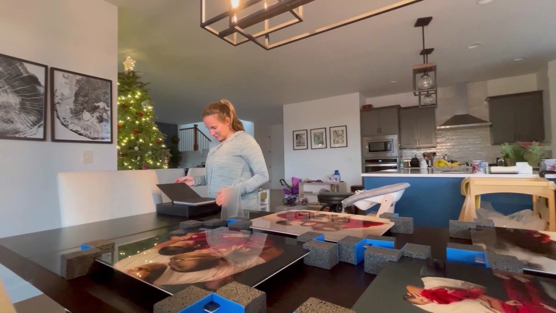 A woman in a light grey hoodie stands at a dining table filled with large, vivid acrylic family portraits protected by blue foam corner guards, while she opens a black heirloom photo box.