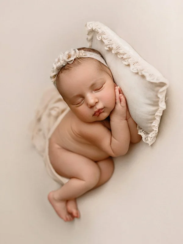 A sleeping newborn baby girl lying on her side, wearing a lace headband, with her hand resting on her cheek and a lacy pillow beside her.
