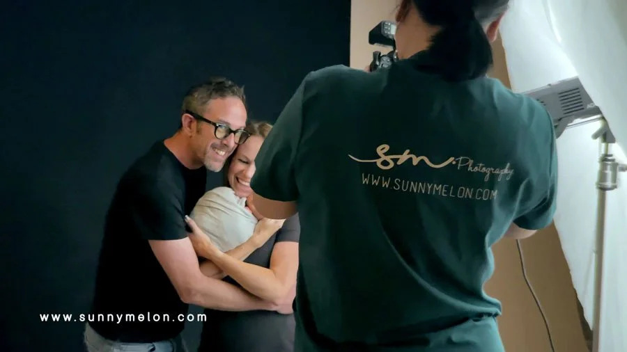 A photographer wearing a "SunnyMelon Photography" shirt captures a candid, emotional moment of a smiling couple hugging while holding their newborn baby in a studio.