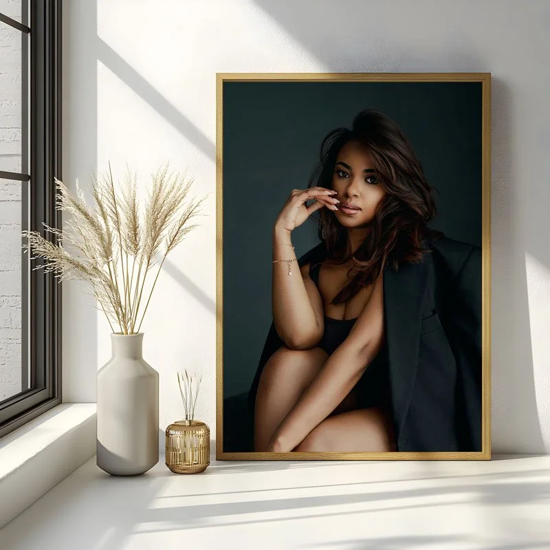 A custom gold-framed fine art portrait of a woman in a black blazer and bodysuit, displayed on a minimalist white sideboard with dried pampas grass and modern decor in a sunlit home.