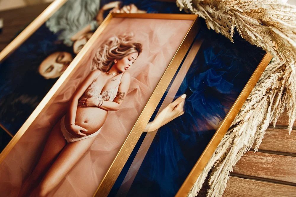 High-end gold-framed fine-art maternity prints displayed on a wooden table with decorative dried grass in a West Linn, Oregon photography studio.