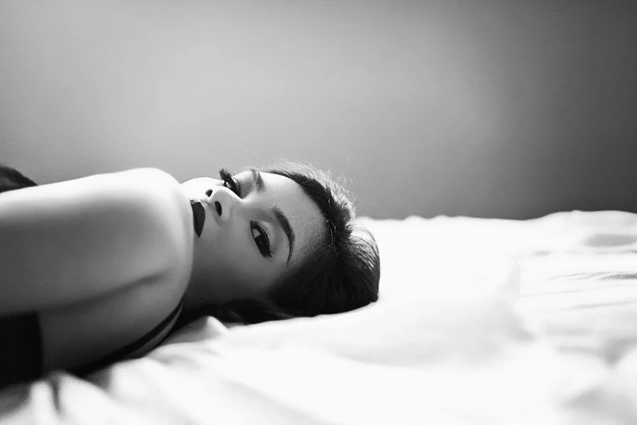 A cinematic black and white close-up of a woman lying on a white bed, looking directly into the camera with dramatic lighting and soft focus.