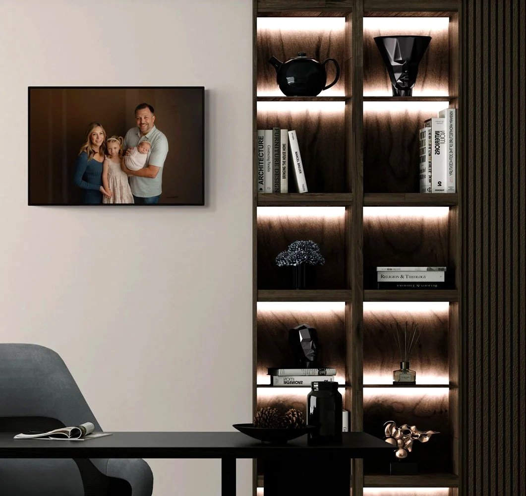 A professional studio family portrait displayed as framed wall art within a luxury home interior in West Linn, Oregon. The portrait features a family of four with a newborn, hanging adjacent to a modern, custom-lit wooden bookshelf.