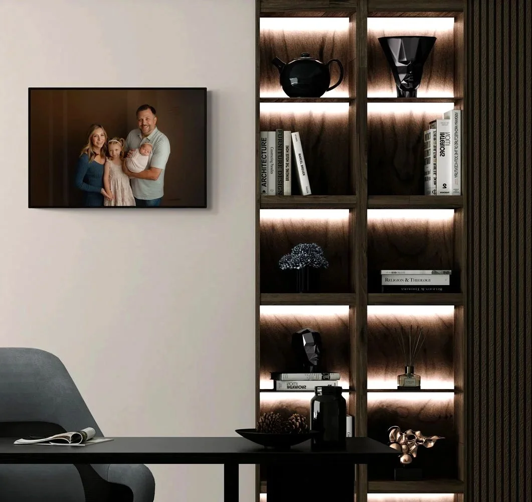 A professional studio family portrait displayed as framed wall art within a luxury home interior in West Linn, Oregon. The portrait features a family of four with a newborn, hanging adjacent to a modern, custom-lit wooden bookshelf.