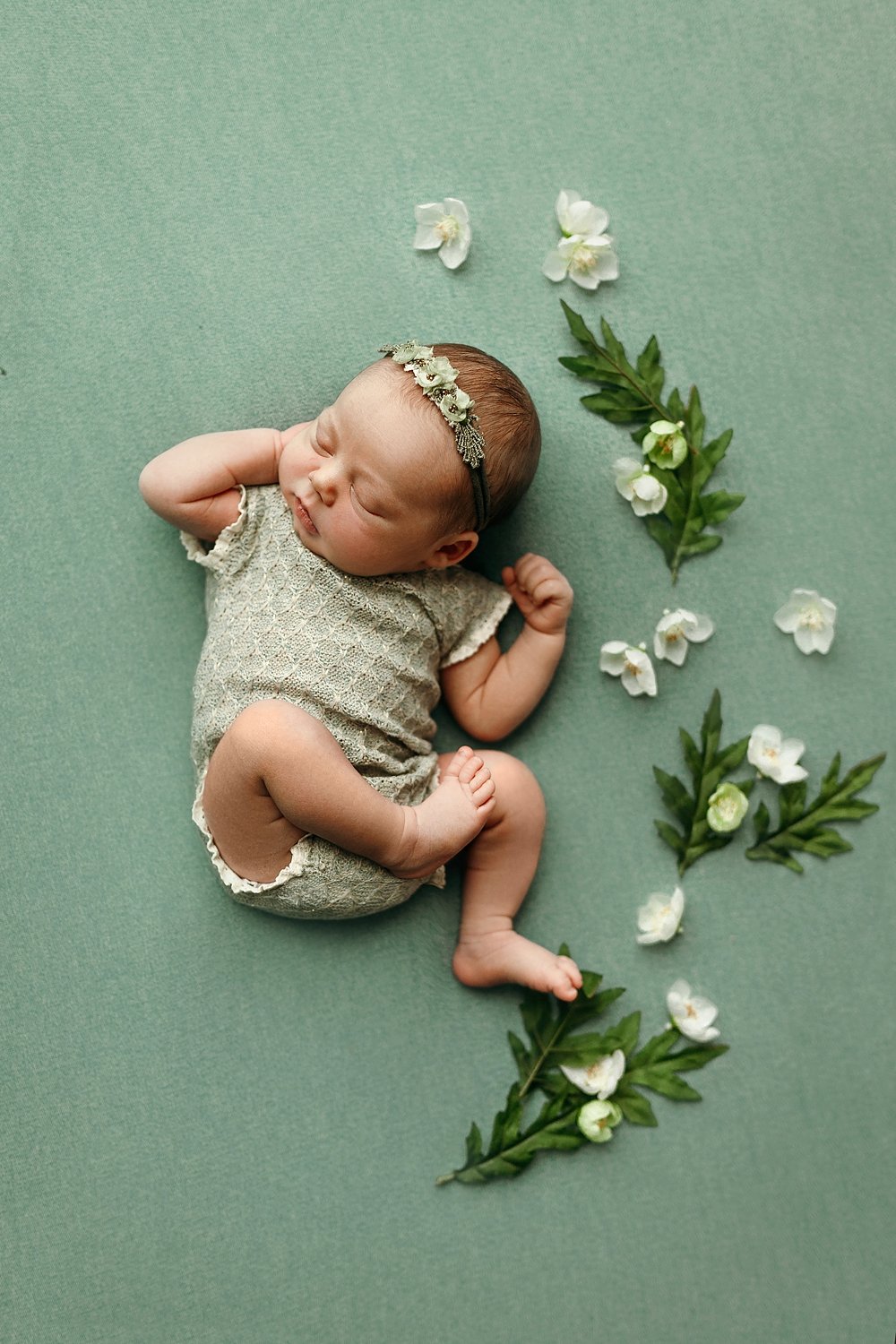 A serene newborn photography setup featuring an infant peacefully sleeping on her back with her hands near her head. The baby is dressed in a light sage green textured knit romper and wears a dainty matching floral headband. She is positioned on a so