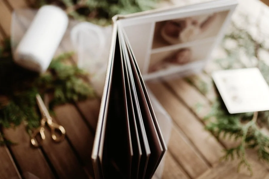 A close-up, artistic view of a thick-paged, custom-designed photography album featuring newborn portraits, resting on a wooden table with gold scissors and greenery accents.
