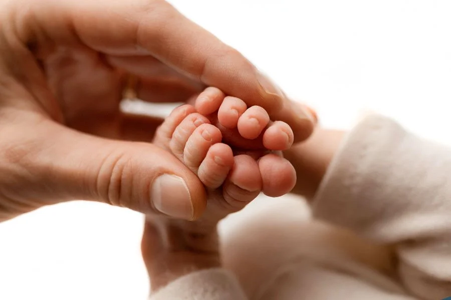A high-detail macro photograph of a newborn baby's tiny toes being gently held by a parent's hand. The image features a clean, bright white background, emphasizing the soft skin and delicate features of the infant. This fine art newborn detail shot c