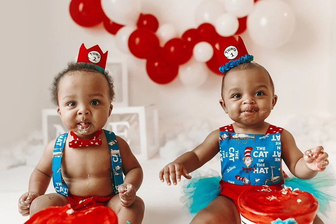 This high-energy twin cake smash portrait captures the delightful aftermath of a first birthday celebration. The twins, dressed in their "Thing 1" and "Thing 2" themed outfits, are shown with frosting-covered faces and hands, having thoroughly enjoye