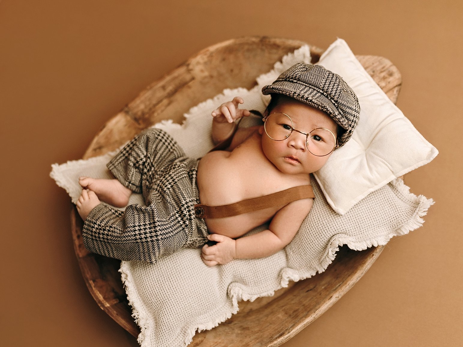 A professional studio newborn portrait by SunnyMelon Photography featuring a baby boy in a vintage academic-inspired pose. The infant is wearing round gold-rimmed glasses, a newsboy cap, and plaid trousers with suspenders. He is posed awake, looking 