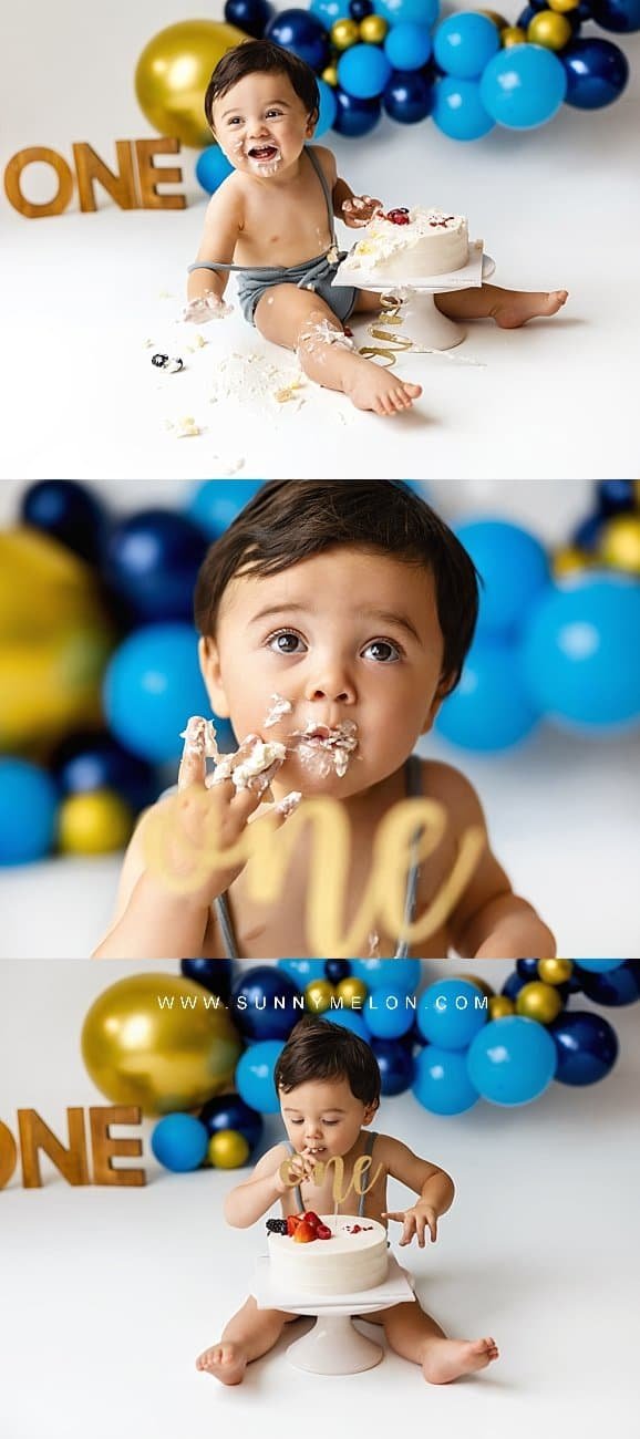 Toddler boy in blue overalls smiling at the camera during a first birthday cake smash photoshoot.