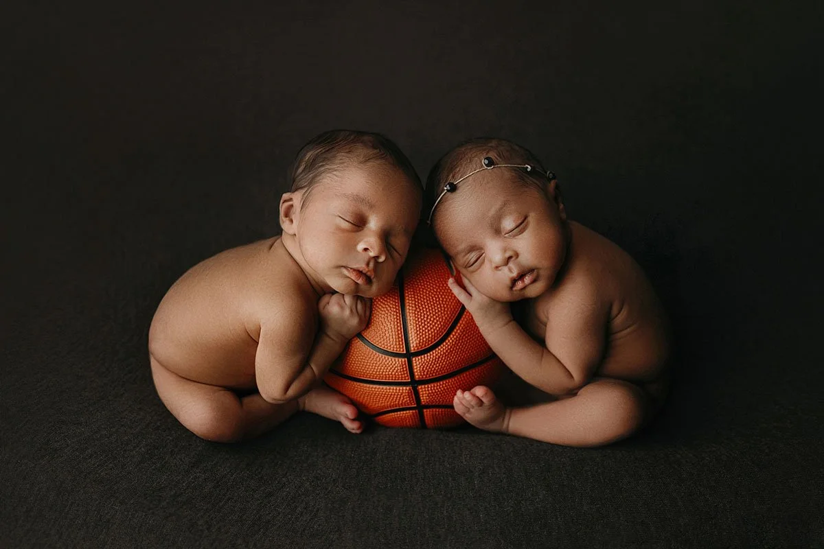 A fine-art studio portrait of the Lillard twins sleeping soundly while resting their heads on a basketball against a dark brown backdrop in West Linn, Oregon. The symmetric, athletic-themed newborn session features soft, focused studio lighting.