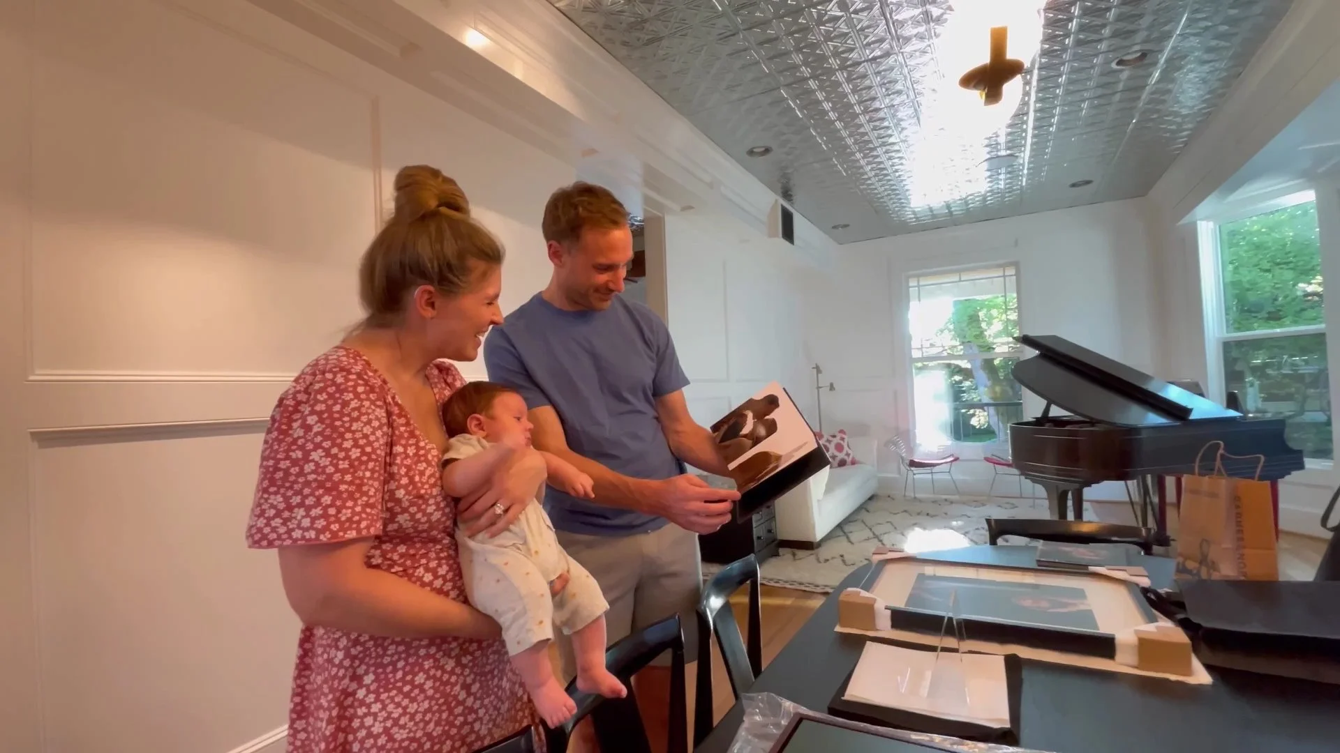 This heartwarming lifestyle image captures new parents sharing a quiet, joyful moment as they flip through a custom-designed newborn photo album. On the table before them sits a professionally framed portrait with protective corners, highlighting a c