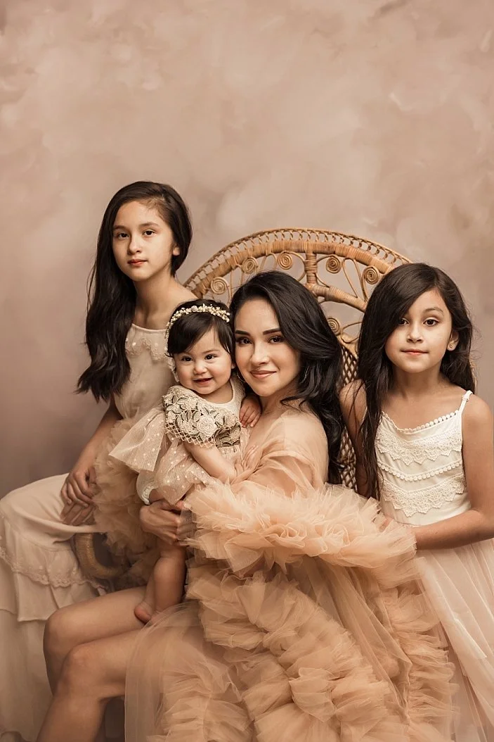 This stunning fine art family portrait captures a mother and her three daughters in a masterfully composed studio setting. The mother, seated in an ornate wicker peacock chair, wears a voluminous peach tulle gown while holding her youngest daughter. 