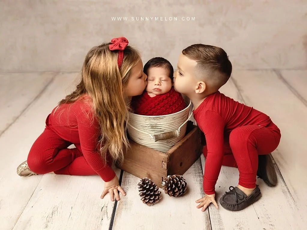 Big sister and brother kissing their newborn sibling in a rustic holiday photography session.