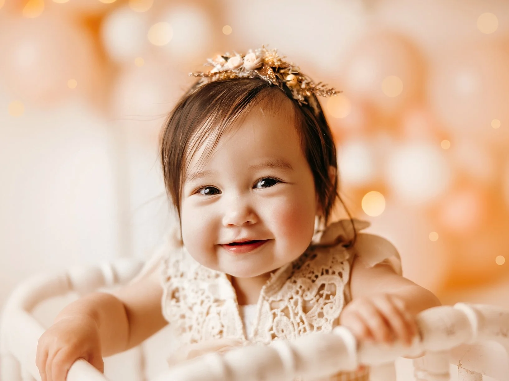 This exquisite infant milestone portrait highlights the soft, ethereal aesthetic of a professional studio session. The baby girl is dressed in a vintage-inspired cream lace romper, topped with a custom floral and gold-leaf headband that adds a touch 