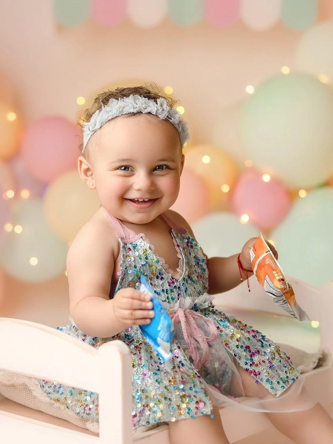This cheerful milestone portrait features a happy toddler girl celebrating her birthday in a whimsical, light-filled studio setting. The subject is dressed in a stunning multi-colored sequin dress with a pink ribbon waist and a matching floral headba