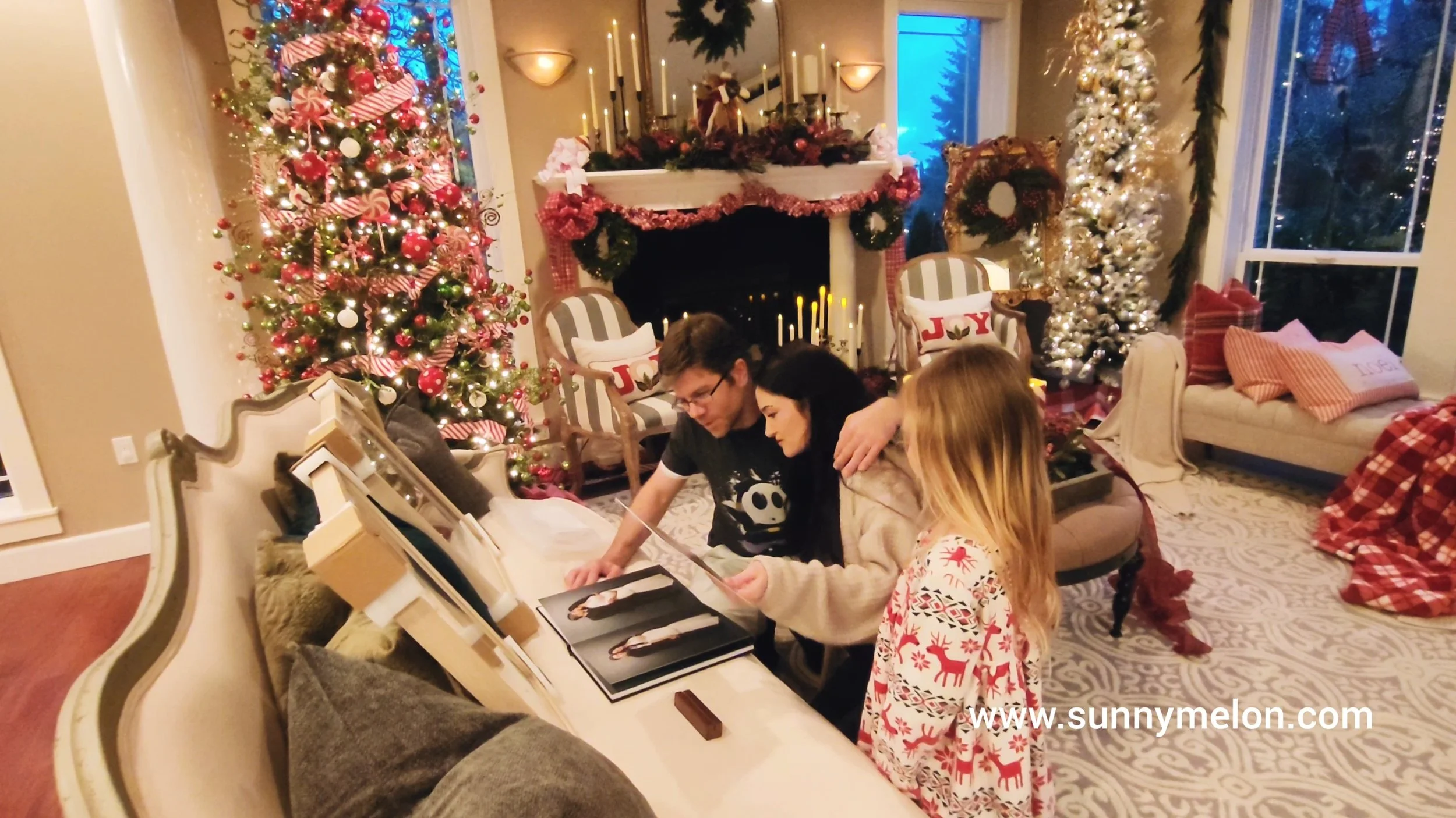 A family of three sits on a couch in a room decorated for Christmas, flipping through a professional photo album while newly framed wall art rests beside them.
