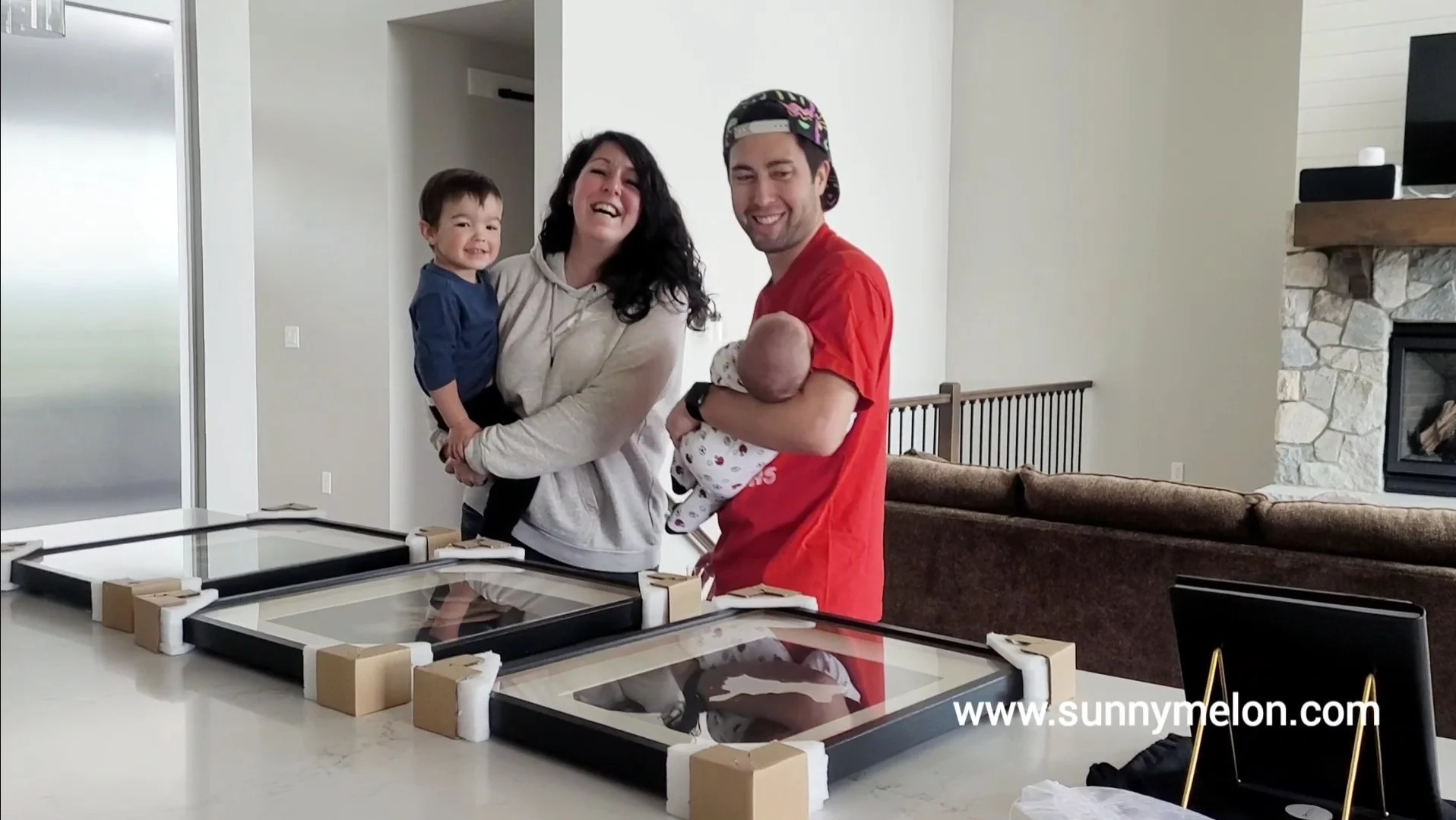 This heartwarming lifestyle image captures a family of four—including a toddler and a newborn—celebrating their professional photography reveal. The parents stand behind a kitchen island displaying three large, high-quality framed portraits featuring