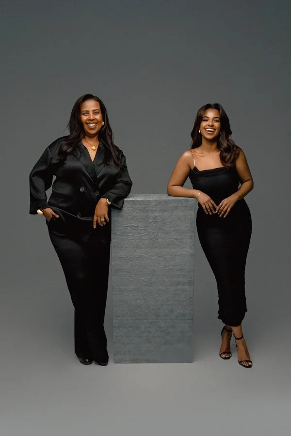 A full-length studio portrait of a mother and daughter laughing, both dressed in elegant black attire and leaning against a grey textured pedestal in front of a minimalist grey backdrop.