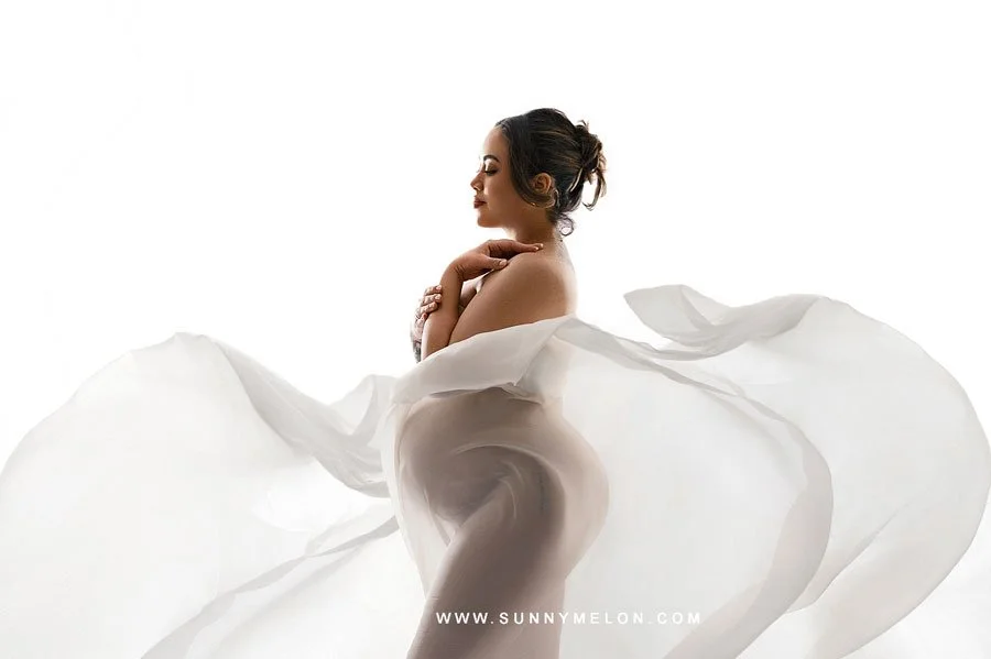 A side-profile maternity portrait of a woman draped in sheer white silk fabric, standing against a bright white backlit background to create a soft, ethereal silhouette.